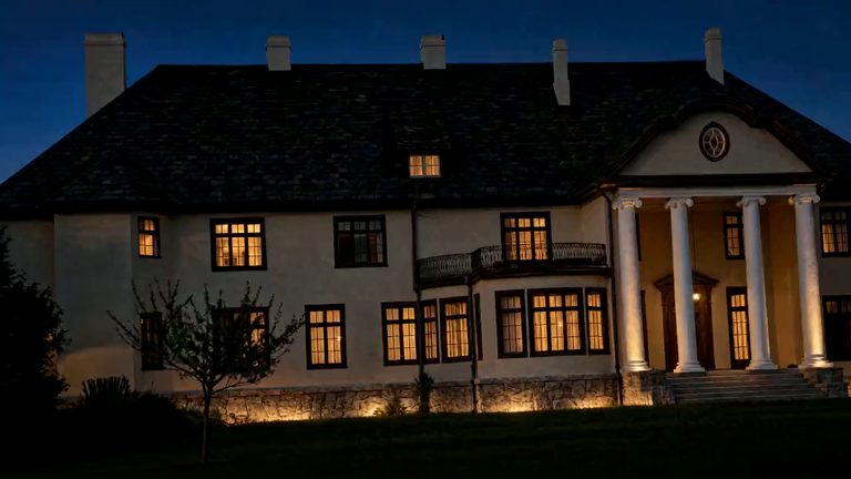 The garden front of the manor house after sunset, seen across the lawn: warm lamplight in the windows, the columns of the portico lit from below, and a deep blue sky above the dark roof