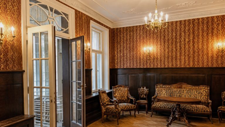 A drawing room with a gilt-framed settee and armchairs under a lit chandelier, a glazed door standing open