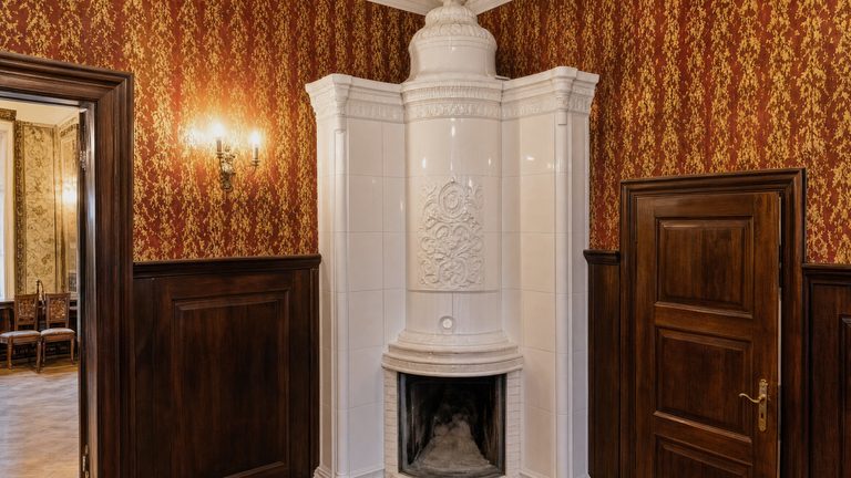 A white tiled corner stove with a moulded crown, between two dark timber doors