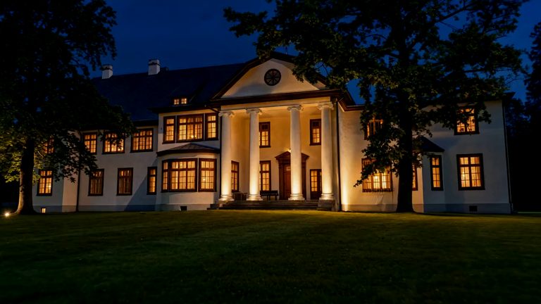 The manor house at dusk seen from the lawn: four white columns under a pediment, warm lamplight in every window, the front door lit under the portico, and mature trees standing either side against a deep blue sky