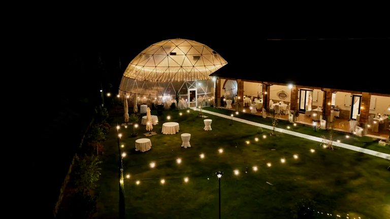 The pavilion at night from the air: a geodesic glass dome lit from within, the long event building beside it, and a lawn strung with lights