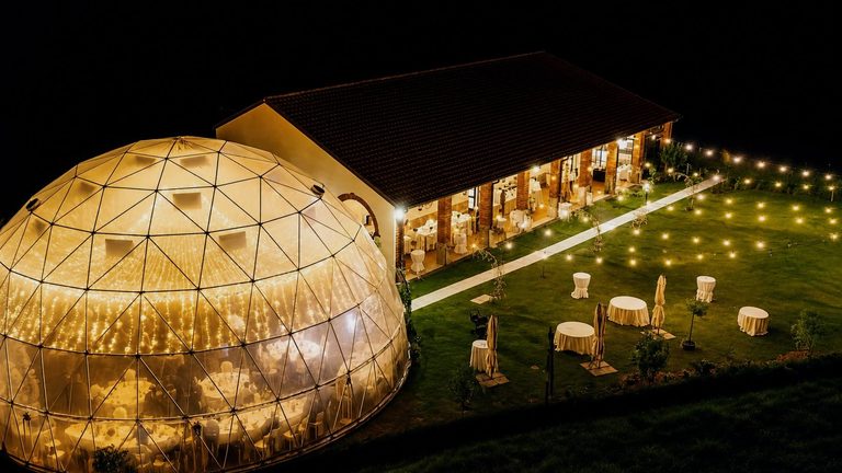 The pavilion and the long event building from the air at night, the lawn between them strung with lights and set with tables