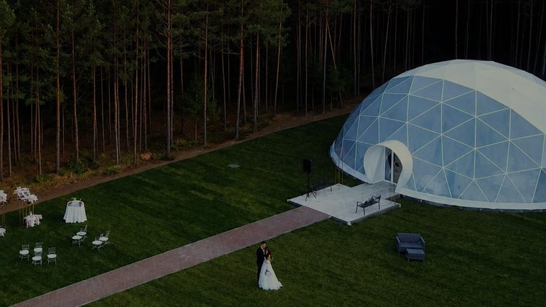 The glass pavilion on the lawn at the edge of a pine wood, a brick path crossing to it and rows of chairs set out on the grass