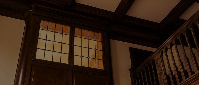 The coffered ceiling and a tall glazed timber screen over the staircase, lit warm from behind, with the carved balustrade beside it