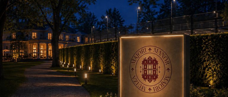 The lamp-lit path to the padel club at blue hour, the clipped yew hedge along it, and the manor house lit beyond