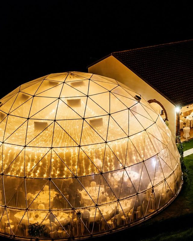 A glass dome lit from inside on a lawn at night, a long building beside it