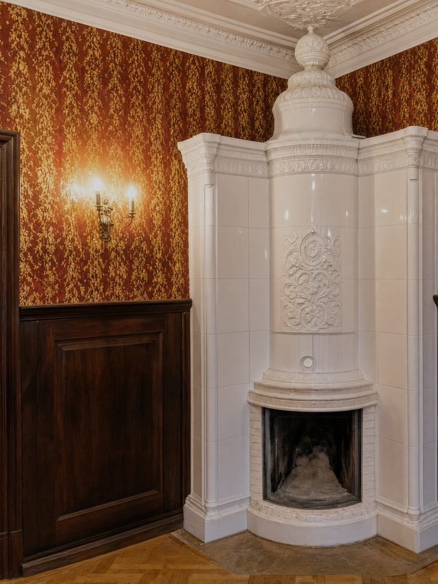 A white tiled stove standing full height in the corner of a room, its firebox open below a moulded crown