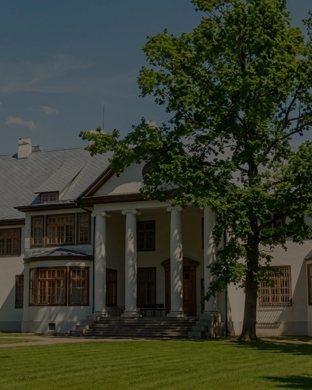 The columned portico of the manor with the tree beside it and the grass in front