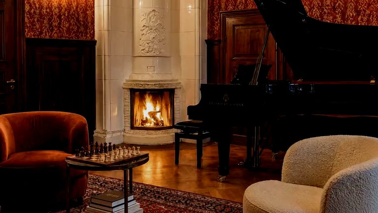 A fire burning in a carved stone chimneypiece in a panelled room, a grand piano beside it and two armchairs drawn up to a chess table