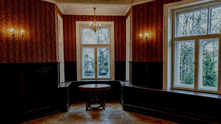 A window bay in a panelled room: two tall windows over a fitted timber seat, sconces lit either side, and the bare trees of the park beyond the glass