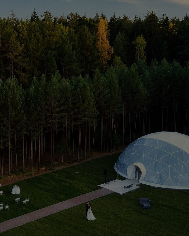 A geodesic glass pavilion on a lawn at the edge of a pine wood, a brick path crossing to it